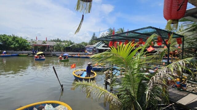 Round boats with people float on a river under a sunny sky in Hoi An, surrounded by palm trees and red lanterns.
