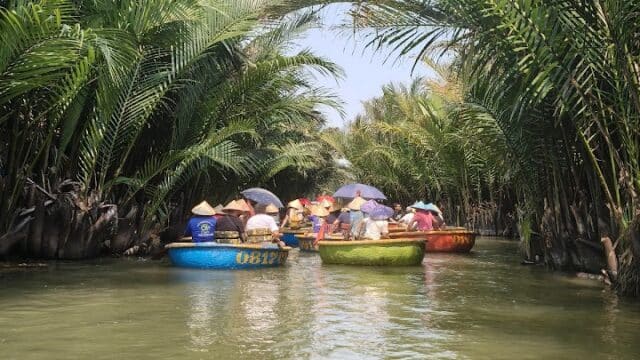 People riding round boats through a river lined with tall palms under Hoi An’s clear blue sky.