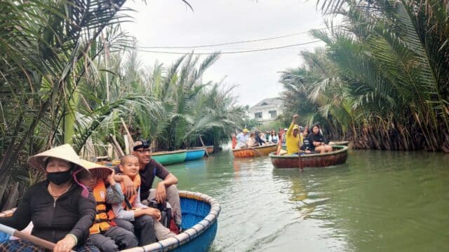 People ride round basket boats in Hoi An through a lush, green waterway lined with palm trees, wearing hats and life jackets.