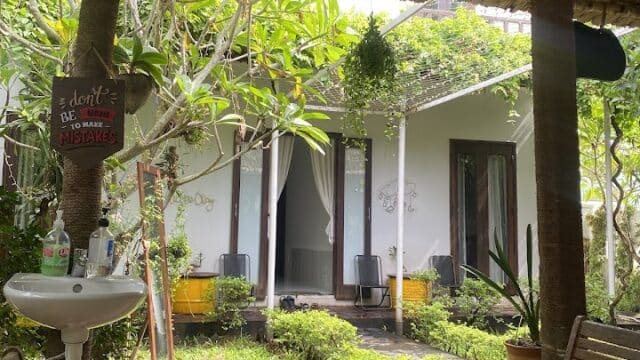 A cozy patio in Hoi An with a glass table, wooden chairs, lush plants, and a white house under a thatched roof.