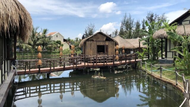Rustic thatched huts and a wooden bridge over a pond with tropical plants, capturing the tranquil charm of Hoi An under a blue sky.