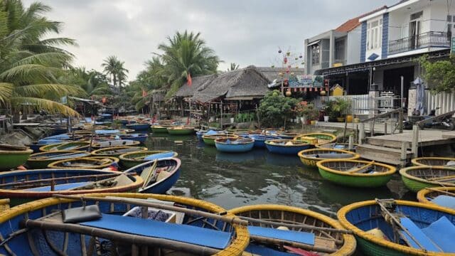 Dozens of round, colorful basket boats float on a canal in Hoi An, lined with palm trees and houses under a cloudy sky.
