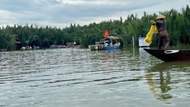 A person casts a yellow fishing net from a boat on Hoi An’s calm river, with palm trees and other boats in the background.