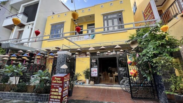 A yellow Hoi An building with lanterns, outdoor plants, a menu sign, and a partially open gate on a sunny day.