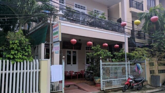 A two-story Hoi An house with red lanterns, a white gate, a scooter, and lush greenery in front.