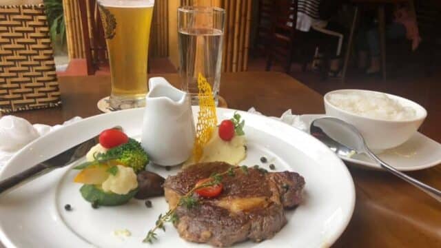 A plate with steak, mashed potatoes, vegetables, and rice awaits on a wooden table in a cozy Hoi An restaurant.