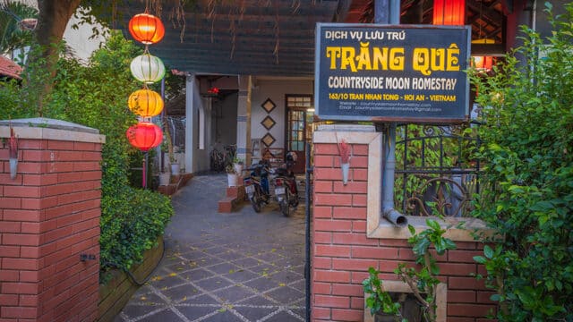 Entrance to Trăng Quê Countryside Moon Homestay in Hoi An, with lanterns, a brick gate, and motorbikes parked in the courtyard.