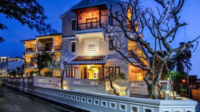 Hoi An hotel with white walls and balconies, warmly lit at dusk, features leafless trees wrapped in glowing string lights out front.