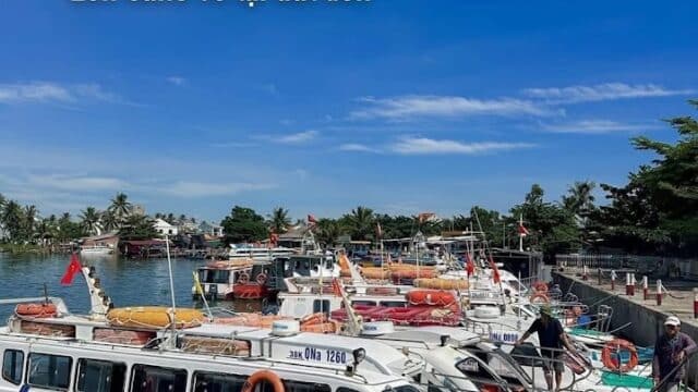 Several white speedboats are docked at a pier in Hoi An under a sunny blue sky; schedule text and Vietnamese writing appear above.
