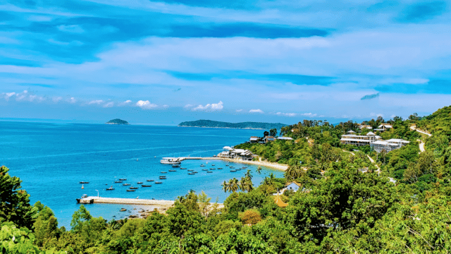 A vibrant coastal view in Hoi An with clear blue water, boats at a small dock, and lush green hills under a bright sky.