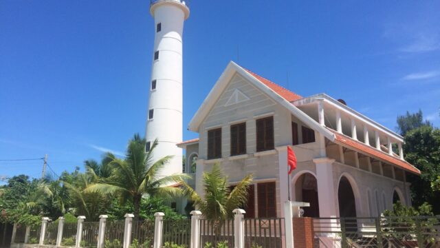White lighthouse and adjacent building with a red roof, palm trees, blue sky, and Hoi An’s coastal charm in the background.