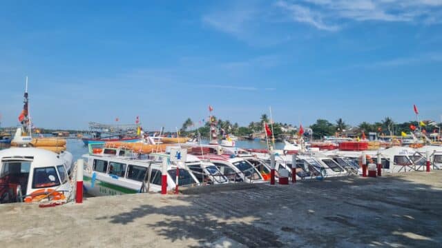 Several colorful boats are docked at a sunny Hoi An marina, with clear blue skies and trees in the background.