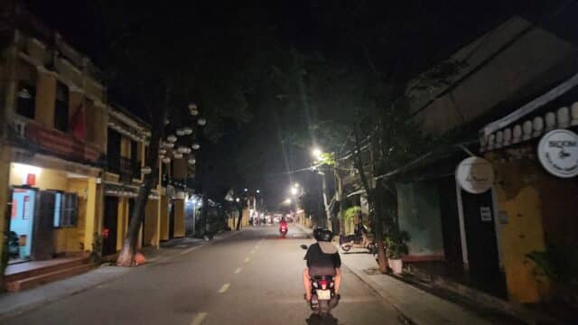 A person rides a scooter down a quiet, dimly lit street lined with buildings in Hoi An at night.