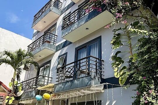 A three-story Hoi An building with balconies, potted plants, string lights, and a pool under a clear blue sky.