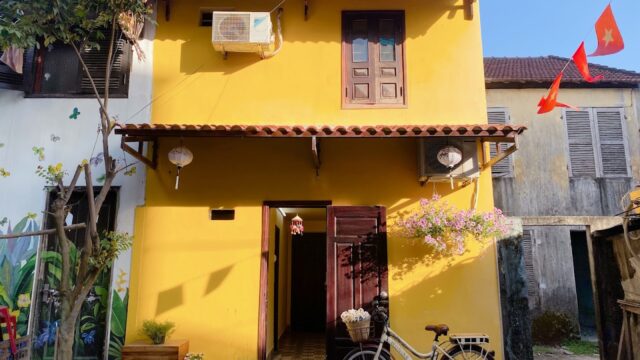 A yellow two-story house in Hoi An with a bike parked outside, flowers, and red flags hanging nearby.