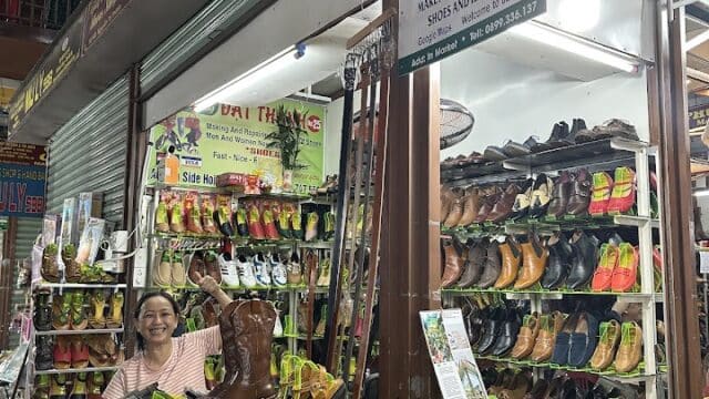 A smiling woman stands in a colorful shoe shop in Hoi An, with various sandals and shoes displayed on vibrant shelves.