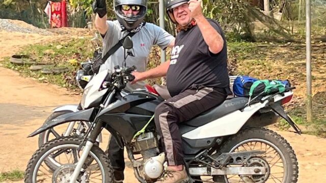 Two people in helmets, one with red horns, pose on a motorcycle outdoors in Hoi An, smiling and gesturing to the camera.