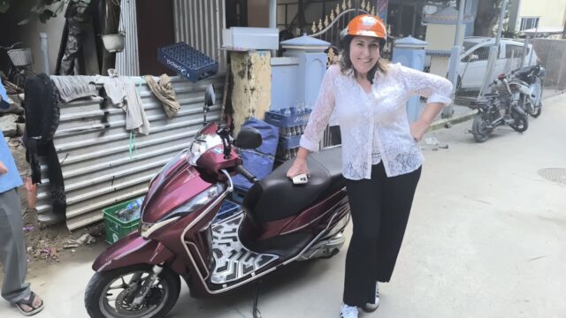 Woman in a white blouse and orange helmet posing with a maroon scooter on a Hoi An street.