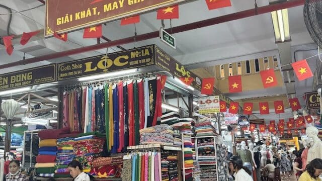 Market scene in Hoi An with fabric stalls, people shopping, and red Vietnamese flags hanging from the ceiling.