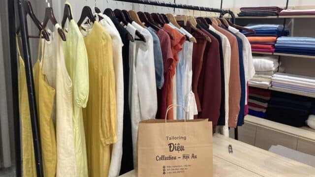 A clothing rack with colorful garments, shelves of folded fabric, and a Tailoring Diệu Hoi An paper bag on a wooden table.