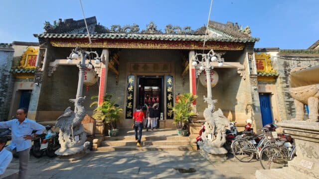 People enter an ornate Chinese temple in Hoi An, with intricate roof details, stone statues, and bicycles parked outside.
