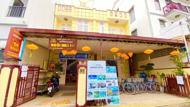 Front view of a Hoi An homestay building with bright lanterns, bicycles, and a booking office display outside.