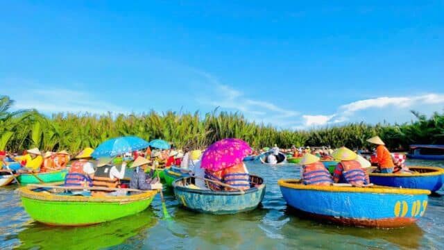 People wearing life vests ride colorful round boats with umbrellas on a river in Hoi An, under a bright blue sky and green trees.