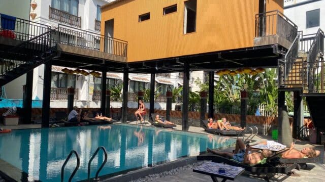 People relaxing by a pool under a modern building with balconies and stairs on a sunny Hoi An day.