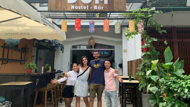 Four people stand smiling with arms around each other in front of DOM Hostel-Bar in Hoi An on a sunny day.