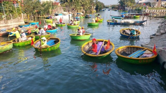 People in round basket boats paddle on a sunny Hoi An river, surrounded by palm trees and houses in the background.