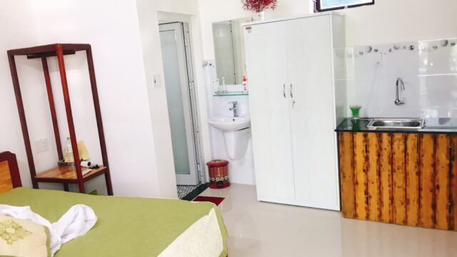 A small, tidy room in Hoi An with a green bed, wardrobe, kitchenette, sink, and an open bathroom featuring a shower stall.