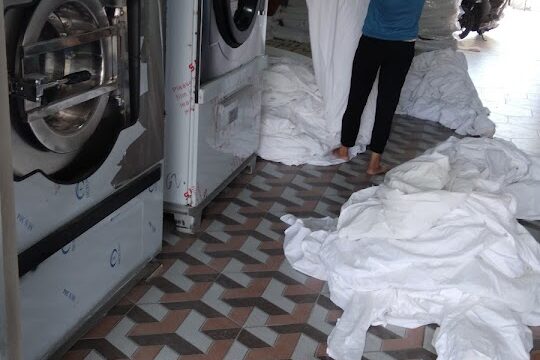 A person folds white sheets next to large washing machines in a Hoi An laundry room with patterned tile flooring.
