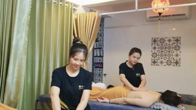 Two women receive back massages from therapists in a Hoi An spa room with green curtains and warm lighting.