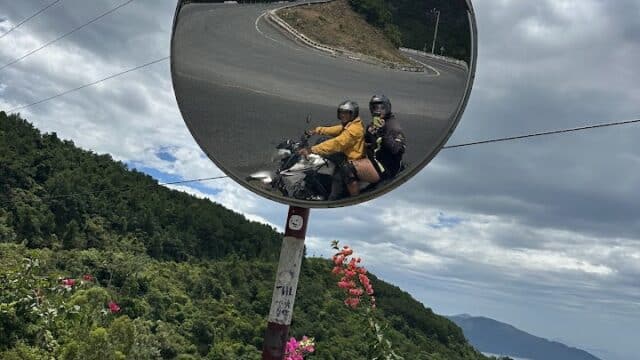 Two people on a motorbike in Hoi An are reflected in a roadside convex mirror with mountains, flowers, and the ocean behind them.