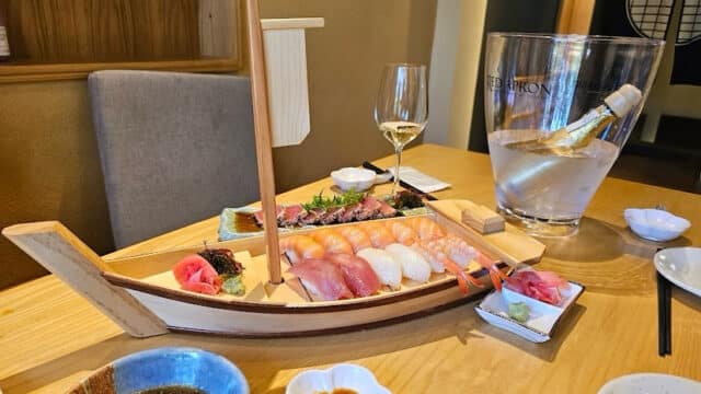 A wooden boat tray with assorted sushi, wine in a bucket, and sauces on a Hoi An restaurant table.