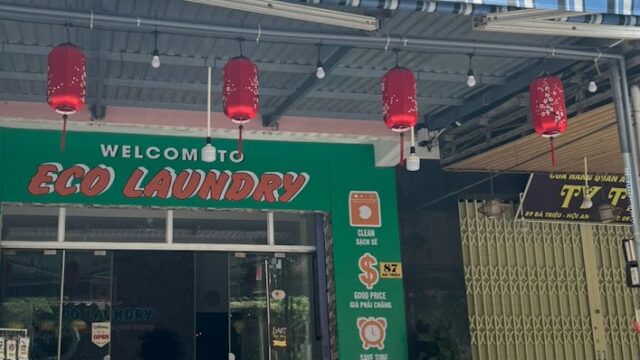 A storefront in Hoi An with a green Eco Laundry sign, red lanterns, and a yellow motorbike parked outside.
