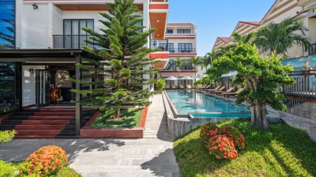 Modern Hoi An hotel with a pool, lounge chairs, green trees, and red flowers under a bright blue sky.