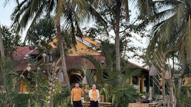 Two people stand on a wooden dock beside a river in Hoi An, surrounded by palm trees and a yellow house in the background.