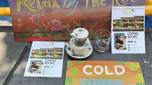 Colorful signs, Hoi An coffee recipe cards, a teacup, glass, and a calendar sit on a table outdoors by a blue pool.