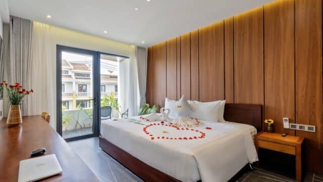 Modern Hoi An hotel room with a double bed, towel swans, rose petals, and stylish wooden panel walls.