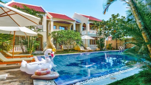 A pool with a large swan float, tropical plants, and a Hoi An villa featuring red roofs and inviting balconies.