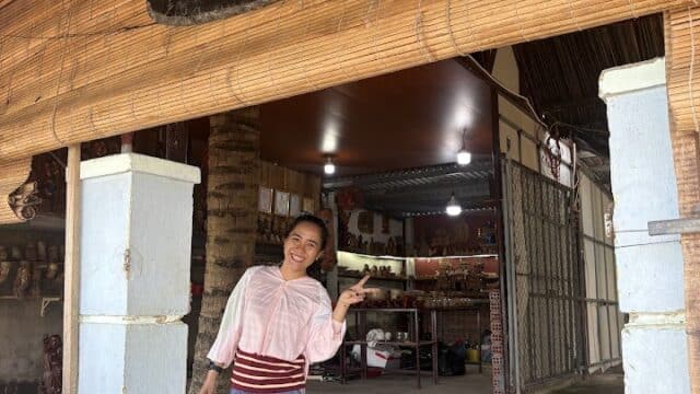 A woman smiles and gestures at the entrance of a Hoi An shop with a wooden sign above the doorway.