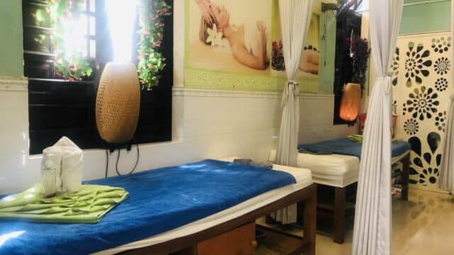 Two massage tables with blue towels in a bright Hoi An spa room, decorated with flowers and a relaxing wall photo.