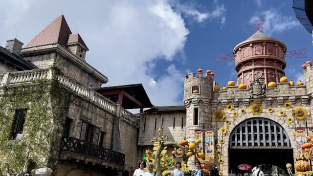 People walk up a cobblestone path toward a decorated castle entrance, reminiscent of Hoi An, under a blue sky with clouds.