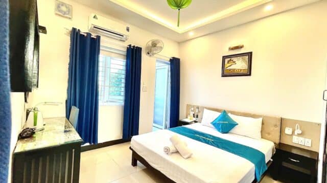 Bright Hoi An hotel room with double bed, blue accents, window, TV, desk, wall art, and neatly placed towels on the bed.
