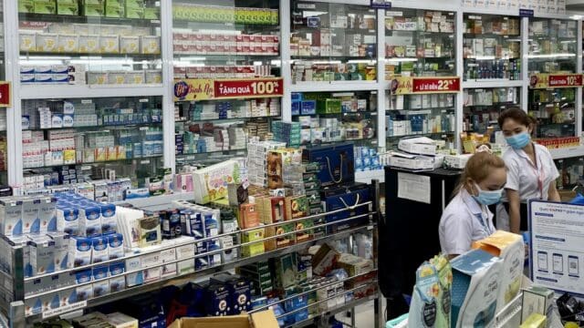Two pharmacy staff in Hoi An wear masks behind a counter surrounded by shelves of medicine and health products.