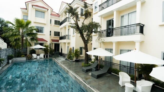 A swimming pool beside a modern cream-colored Hoi An apartment building with balconies and poolside lounge chairs.