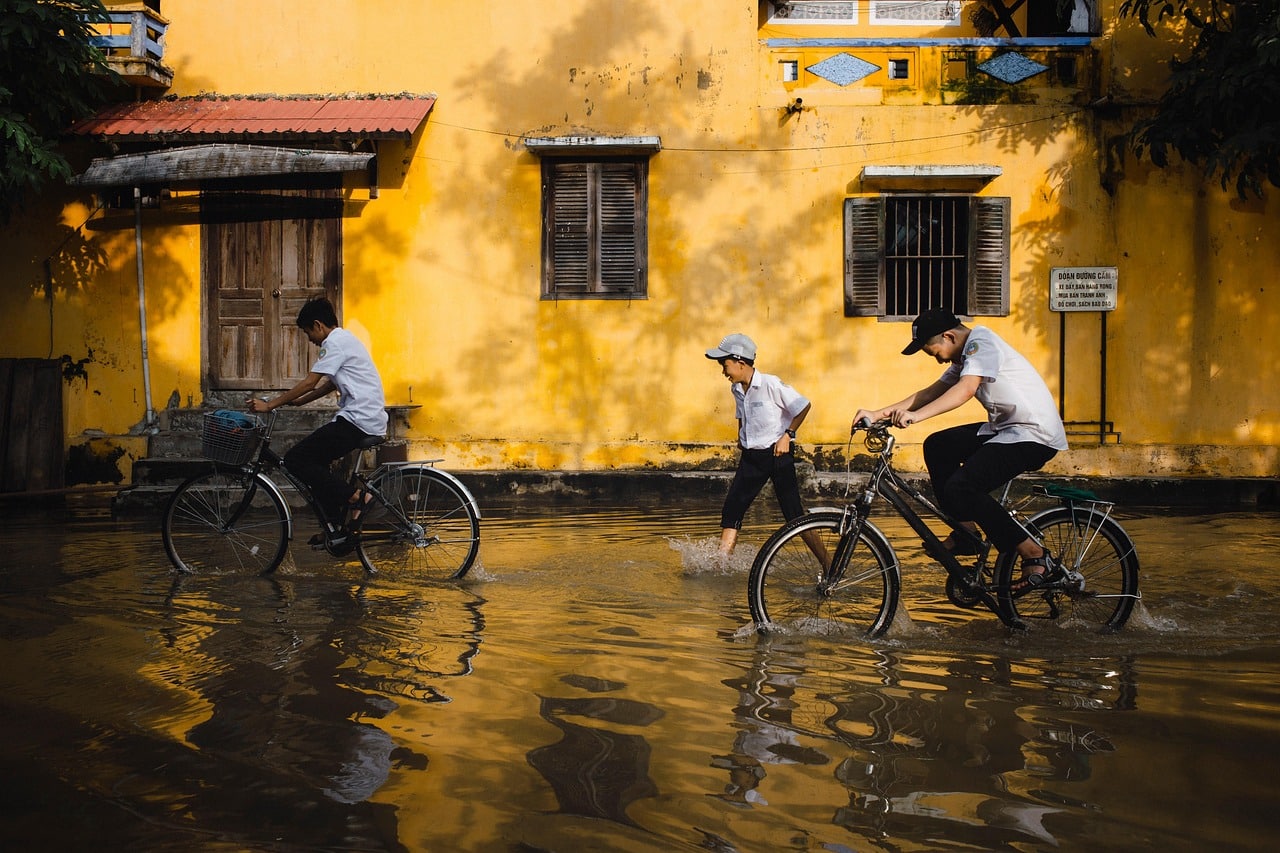 Schoolchildren pushing bicycles through shallow floodwater in an old Vietnamese town