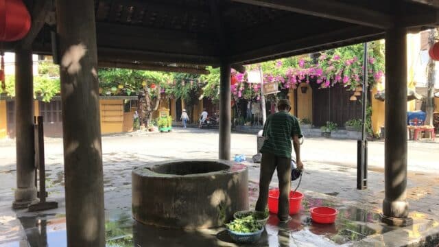 A man washes vegetables by an old well under a roofed structure in a sunlit, flower-lined street of Hoi An.
