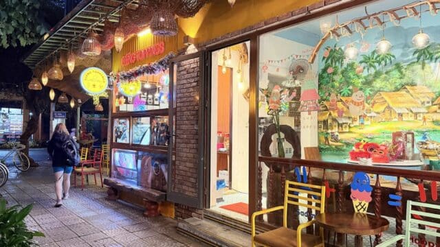 A woman strolls past a brightly lit Hoi An ice cream shop with colorful chairs and a mural on the front window at dusk.